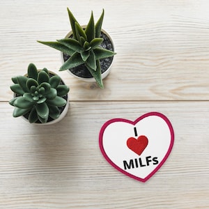 May include: A heart-shaped patch with a red border and the words "I ❤️ MILFs" embroidered on it. Two small potted succulents are also visible on a light wood grain surface.