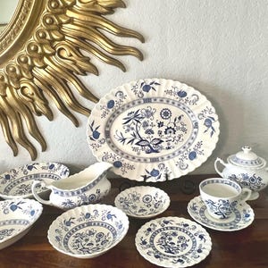 May include: A collection of blue and white porcelain tableware, including plates, bowls, a teapot, and a creamer. The pieces feature a floral design and scalloped edges. A large gold sunburst mirror is in the background.