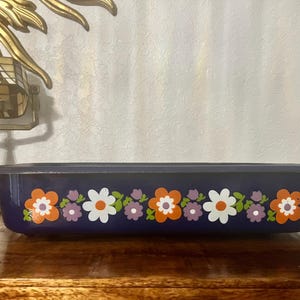 May include: A rectangular, navy blue enamel baking dish with a floral pattern. The flowers are orange, white, and purple with green leaves. The dish sits on a wooden surface.