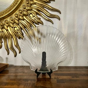 May include: A clear, glass seashell-shaped plate displayed on a black stand. The plate has a ribbed texture and is set against a backdrop of a gold sunburst mirror and a wooden surface.