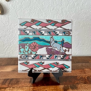 May include: Square ceramic tile with a Southwestern design. The tile features a howling wolf, cacti, and geometric patterns in shades of teal, pink, purple, and white. The tile is displayed on a black stand.
