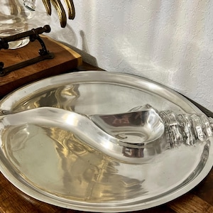 May include: A silver-colored serving platter with a seashell design. The platter has a large oval shape with a built-in dip bowl. The seashell detail is on the right side of the platter.