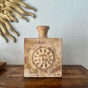 May include: A square, textured vase with a sun face design. The vase is a mix of beige, brown, and red hues, with a cylindrical neck. The sun design is in the center of the vase. The vase is on a wooden surface.