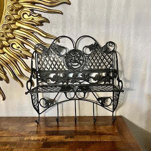 May include: Black metal bench-style wall shelf with decorative floral and scrollwork details. Features a diamond-patterned back and three hooks below. The shelf is displayed on a wooden surface.