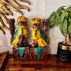May include: Two yellow and green ceramic Foo Dog statues with brown accents. Each statue sits atop a brown base with teal accents. A gold sunburst wall decor and a potted plant are in the background.
