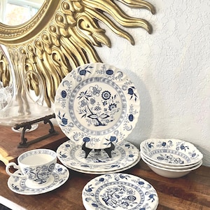 May include: A collection of white ceramic dinnerware with a blue floral pattern. The set includes plates, bowls, and a teacup with saucer. The plates have scalloped edges. The teacup and saucer are on the left side of the image.
