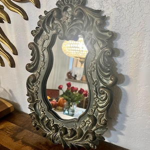 May include: An ornate, oval-shaped mirror with a dark bronze finish. The frame features intricate floral and scrollwork details. The mirror reflects a room with a chandelier and a vase of red roses.