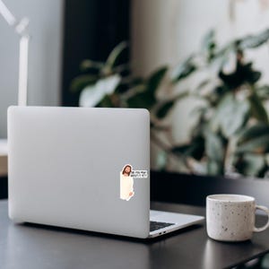 Op de afbeelding: Een laptop met een sticker van Jezus met de tekst "DID YOU PRAY ABOUT IT YET?" staat op een donkere ondergrond. Rechts van de laptop staat een witte, gespikkelde mok. Groene planten staan op de achtergrond.