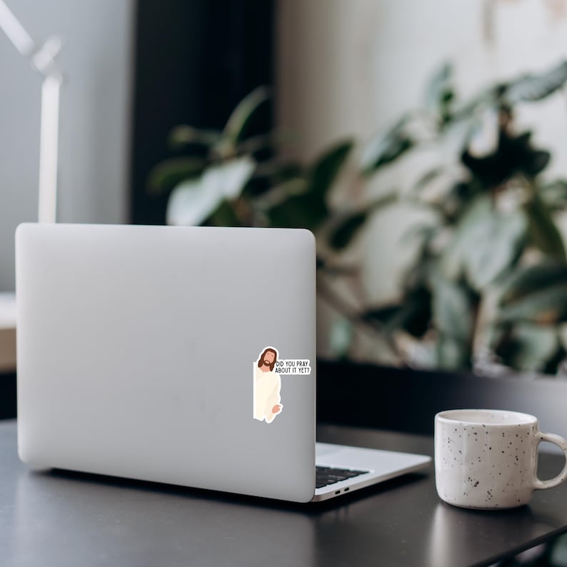 Op de afbeelding: Een laptop met een sticker van Jezus met de tekst "DID YOU PRAY ABOUT IT YET?" staat op een donkere ondergrond. Rechts van de laptop staat een witte, gespikkelde mok. Groene planten staan op de achtergrond.
