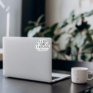 May include: A white laptop with a sticker that reads "GOD FAMILY COUNTRY in that order" in black script. A speckled white mug sits to the right of the laptop. The scene is set on a dark surface with greenery in the background.