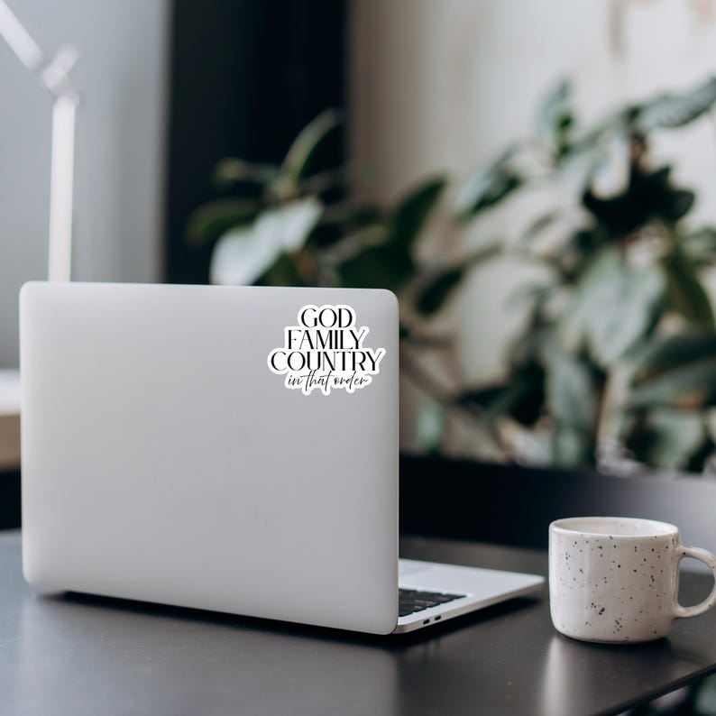 May include: A white laptop with a sticker that reads "GOD FAMILY COUNTRY in that order" in black script. A speckled white mug sits to the right of the laptop. The scene is set on a dark surface with greenery in the background.