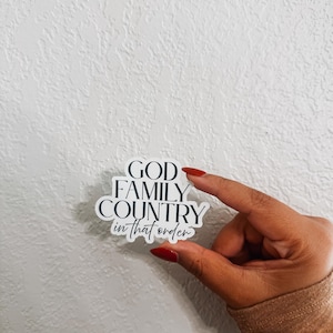 May include: A white sticker with black text that reads "GOD FAMILY COUNTRY in that order." The sticker is held between two fingers with red nail polish against a textured white background.