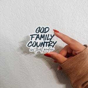 May include: A white sticker with the text "GOD FAMILY COUNTRY in that order" in a black, bold font. The sticker is held by a hand with red nail polish against a textured white background.