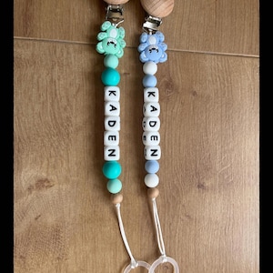 Op de afbeelding: Twee gepersonaliseerde fopspeenclips voor baby's. Eén clip is lichtblauw met een blauwe beer, de andere is mintgroen met een groene beer. Elke clip spelt "KADEN" met witte letterkralen. De clips hebben houten accenten.
