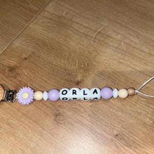 May include: A baby's dummy clip with the name "ORLA" spelled out in white letter beads. The clip has a wooden clip, a lavender daisy, and lavender and beige beads. A white string is attached.