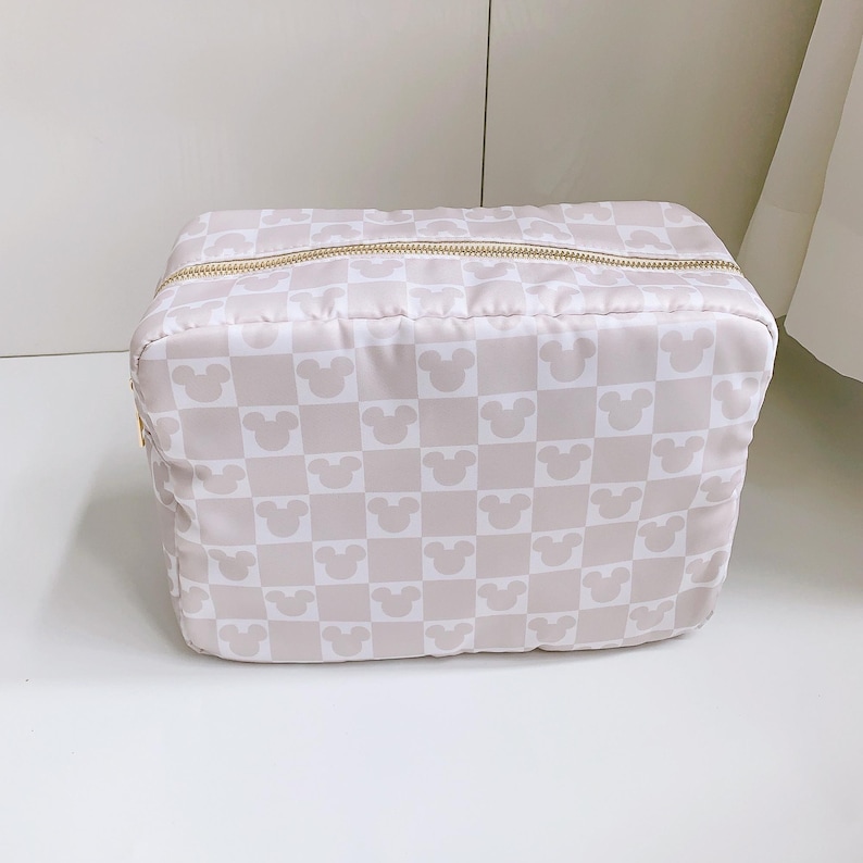 Op de afbeelding: Een beige en witte make-uptas met een dambordpatroon van Mickey Mouse-silhouetten. De tas heeft een gouden ritssluiting en een rechthoekige vorm. De tas staat op een wit oppervlak.