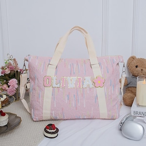 Peut inclure: Sac fourre-tout rose avec des poignées et une bandoulière crème. Le sac porte le nom "OLIVIA" en lettres dorées à paillettes et une fleur rose. Le sac présente un motif texturé avec des lignes verticales aux couleurs pastel.