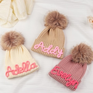 Puede incluir: Tres gorros de punto con pompones de piel sintética. Uno es de color crema con el nombre "Adella" en escritura rosa, otro es beige con "Addy", y el tercero es rosa con "Evelyn". Los gorros están sobre una superficie blanca.