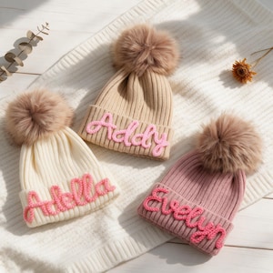 Puede incluir: Tres gorros de punto acanalado con grandes pompones de piel sintética. Un gorro beige lleva el nombre Addy en rosa, un gorro crema lleva el nombre Adella en rosa y un gorro rosa empolvado lleva el nombre Evelyn bordado en rosa.
