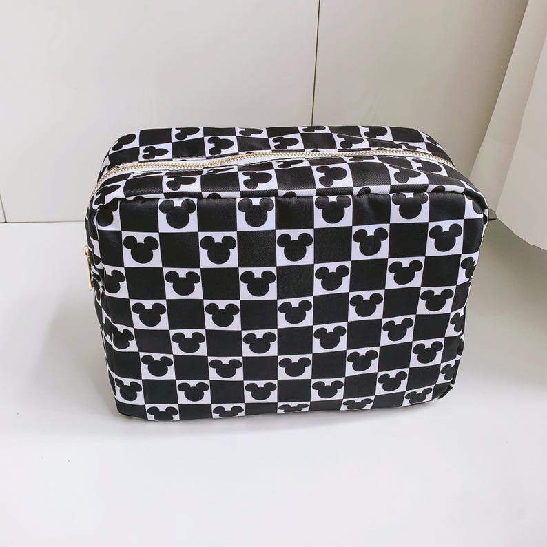 Op de afbeelding: Zwart-wit geruite make-uptas met Mickey Mouse-silhouetten. De tas heeft een rechthoekige vorm met een ritssluiting en een goudkleurige ritssluiting. De tas is gemaakt van een glanzend materiaal.