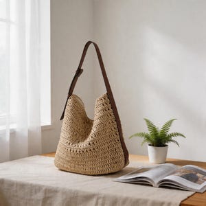 Op de afbeelding: Een beige geweven schoudertas met een bruine leren riem. De tas is gemaakt van een textuurrijk, open geweven materiaal. De riem is met een gesp aan de tas bevestigd. De tas staat op een tafel met een boek en een kleine potplant.