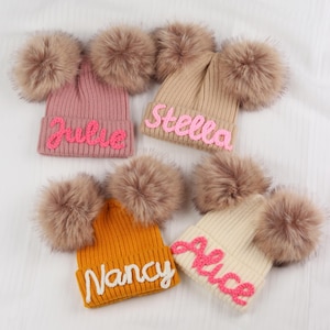 May include: Five knitted beanies in various colors: pink, tan, mustard yellow, and cream. Each beanie features a fluffy pom-pom on each side and a name embroidered across the front in a contrasting color. Names include Julie, Stella, Nancy, and Alice.
