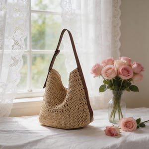 Op de afbeelding: Een beige geweven schoudertas met een bruine leren riem. De tas staat op een wit oppervlak naast een vaas met roze rozen. De tas heeft een grote opening en een afgeronde vorm.