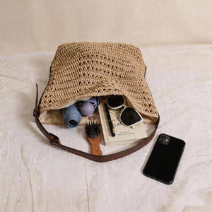 Op de afbeelding: Een beige geweven handtas met een bruine leren riem, open om de inhoud te onthullen, waaronder een zonnebril, een boek, een pen, een haarborstel, een opgerolde blauwe paraplu en een smartphone.