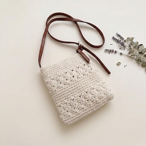 May include: A cream-colored crochet crossbody bag featuring two horizontal bands of floral-inspired openwork patterns. The bag includes a thin, dark brown faux leather strap attached with a silver-toned metal ring and a small brown leather tab.