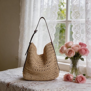 Op de afbeelding: Een beige geweven draagtas met bruine leren bandjes. De tas heeft een grote open bovenkant en een afgeronde bodem. De tas staat op een wit kanten tafelkleed, naast een vaas met roze rozen.