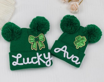 May include: Two green knit beanies with large pom-poms. One beanie has the word "Lucky" in white script and a green bow. The other beanie has the name "Ava" in white script and a sequined shamrock. Perfect for St. Patrick's Day.