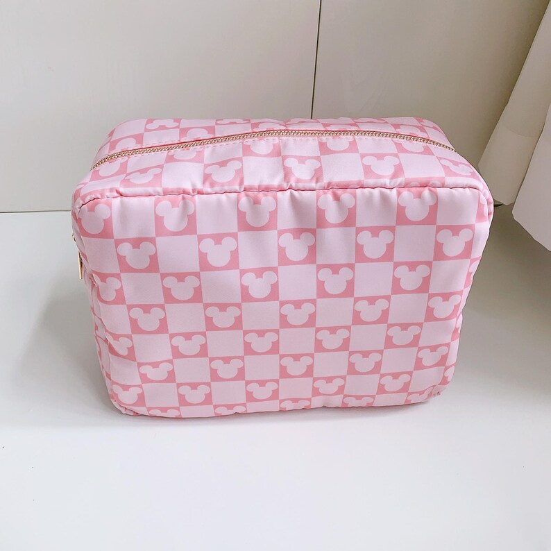 Op de afbeelding: Roze en witte geruite cosmetische tas met een gouden rits. De tas heeft een patroon van Mickey Mouse-hoofden in een dambordpatroon. De tas is rechthoekig en lijkt gemaakt van een zacht, gewatteerd materiaal.