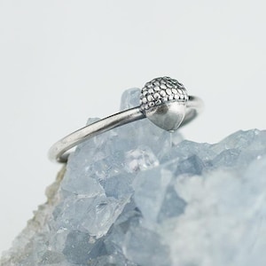 May include: A silver ring with an acorn design. The ring is on a blue crystal surface.