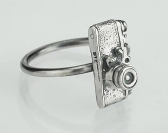 Puede incluir: Un anillo de plata con un dije de cámara detallado. El dije de la cámara tiene una superficie texturizada y una lente con un borde negro.