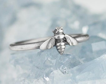 May include: A silver ring with a bee design. The bee is detailed with wings, legs, and a stinger. The ring is set against a blue background.