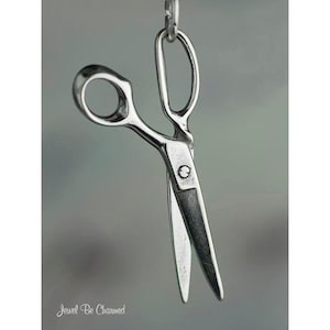 May include: A silver-colored pair of scissors suspended from a chain. The scissors are open, with sharp blades and a classic design. The image has a soft focus background and the text "Jewel Be Charmed" is visible at the bottom.