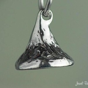 May include: A close-up of a silver-colored charm in the shape of a Hershey's Kiss. The charm has a textured surface and a loop at the top for attaching to a necklace or bracelet. The charm is suspended against a soft, blurred background.