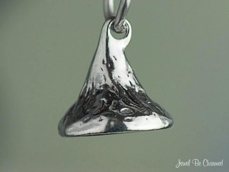 May include: A close-up of a silver-colored charm in the shape of a Hershey's Kiss. The charm has a textured surface and a loop at the top for attaching to a necklace or bracelet. The charm is suspended against a soft, blurred background.