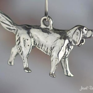 May include: Silver charm of a hound dog in a running pose. The charm is detailed with a textured surface and has a loop at the top for attaching to a bracelet or necklace.