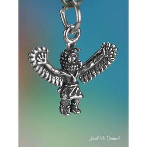 May include: A silver-colored charm in the shape of a Native American figure with outstretched arms and detailed wings. The charm is suspended from a small metal ring. The figure is wearing a detailed headdress and clothing. The background is a gradient of blue and green.