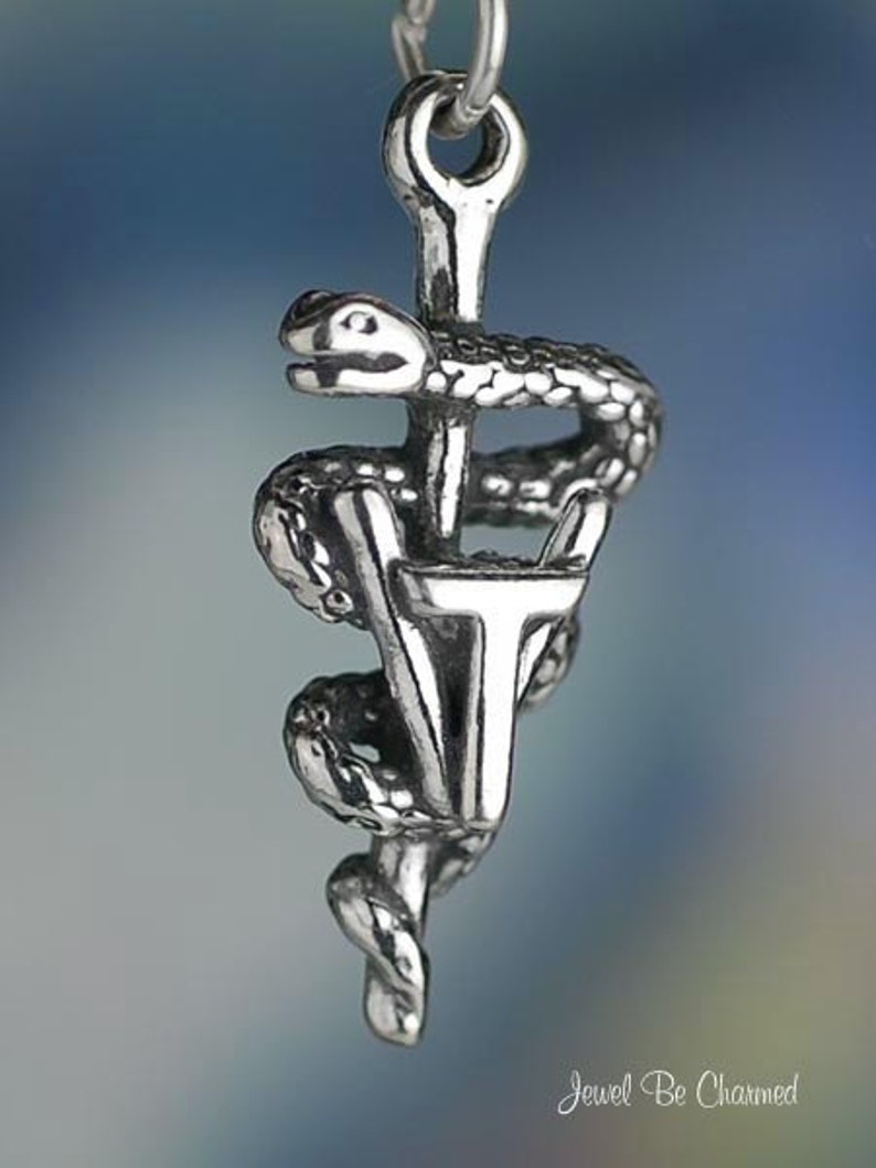 May include: A silver-toned pendant featuring the Rod of Asclepius, a symbol of medicine. The design includes a snake coiled around a staff, with the letters "VT" at the base. The pendant is suspended from a chain.