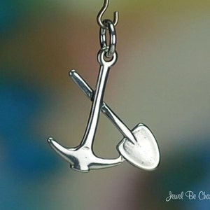 May include: Silver charm featuring a small anchor and a shovel, both crossed. The charm is designed to be attached to a bracelet or necklace.