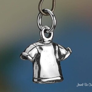 May include: A small, silver-colored charm in the shape of a t-shirt, suspended from a metal ring. The charm has detailed features, including sleeves and a neckline. The background is a gradient of blue and gray. The text "Jewel Be Charmed" is in the bottom right corner.