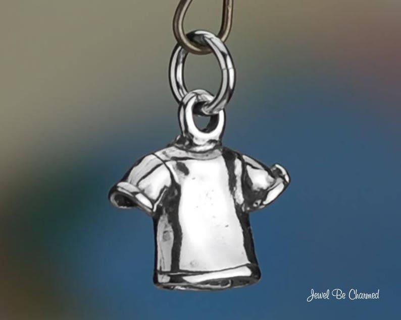 May include: A small, silver-colored charm in the shape of a t-shirt, suspended from a metal ring. The charm has detailed features, including sleeves and a neckline. The background is a gradient of blue and gray. The text "Jewel Be Charmed" is in the bottom right corner.