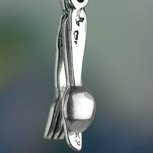 Może przedstawiać: Zbliżenie srebrnego charmsa z motywem widelca i łyżki. Charms zawieszony jest na małym k&oacute;łeczku. Tło to rozmyta mieszanka niebieskiego i zielonego. Charms ma błyszczące wykończenie.