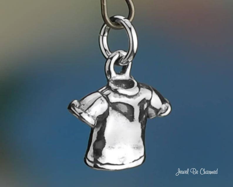 May include: A small, silver-colored charm in the shape of a t-shirt, suspended from a metal ring. The charm has detailed features, including sleeves and a neckline. The background is a gradient of blue and green.