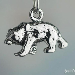 May include: A silver charm in the shape of a bear walking. The charm is detailed with a textured surface and has a loop at the top for attaching to a bracelet or necklace.