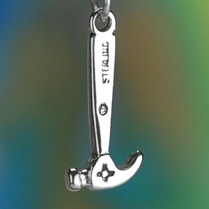 May include: A silver-colored hammer charm with the word "STERLING" engraved on the handle. The hammer head features a star-shaped design. The charm is suspended from a silver chain against a blurred, colorful background.
