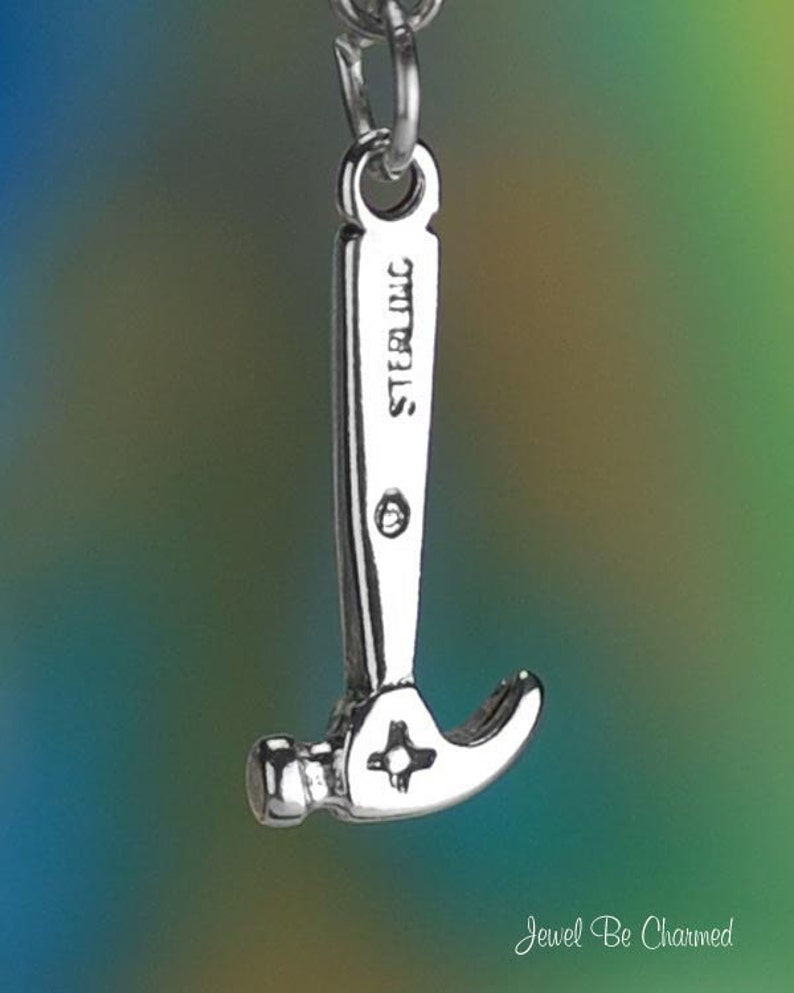 May include: A silver-colored hammer charm with the word "STERLING" engraved on the handle. The hammer head features a star-shaped design. The charm is suspended from a silver chain against a blurred, colorful background.