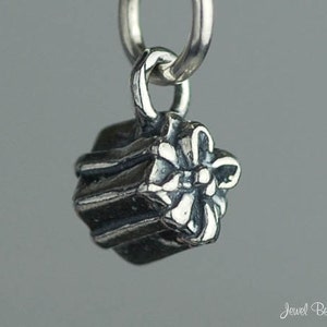 May include: A small, silver-toned charm in the shape of a wrapped gift with a bow. The charm is suspended from a silver ring, and the gift has textured, ribbed sides. The background is a solid, light gray.